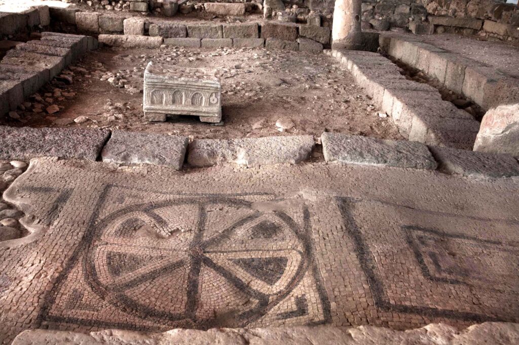 Interior of the first-century Magdala synagogue with geometric mosaic flooring, stone benches, and a replica of the Magdala Stone.