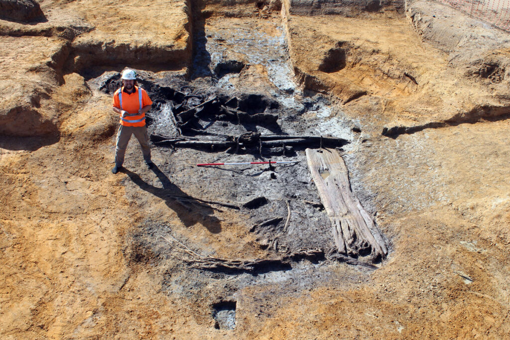 Bronze Age waterhole in Gloucester with preserved wooden structures and an archaeologist standing beside the excavation.