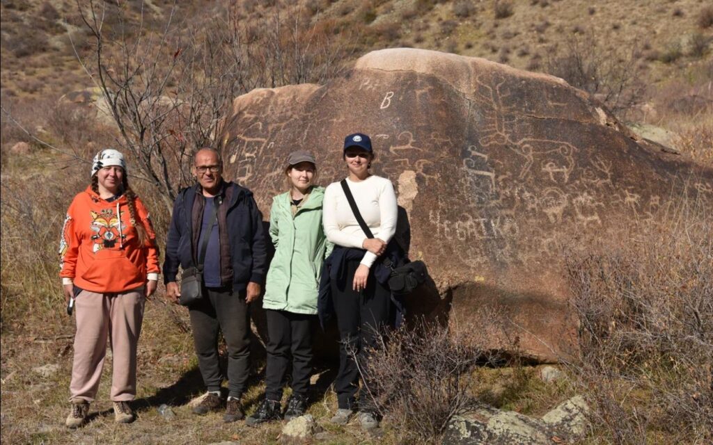 Archaeological expedition members standing beside a large rock with ancient petroglyphs in Kazakhstan’s Koksu River Valley, originally sketched by Pyotr Semyonov-Tyan-Shansky in 1856.