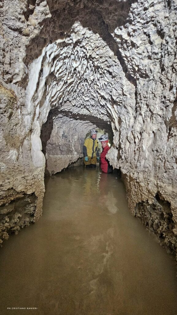 Two researchers wearing helmets and protective clothing stand in water inside an ancient rock-cut aqueduct in central Italy.