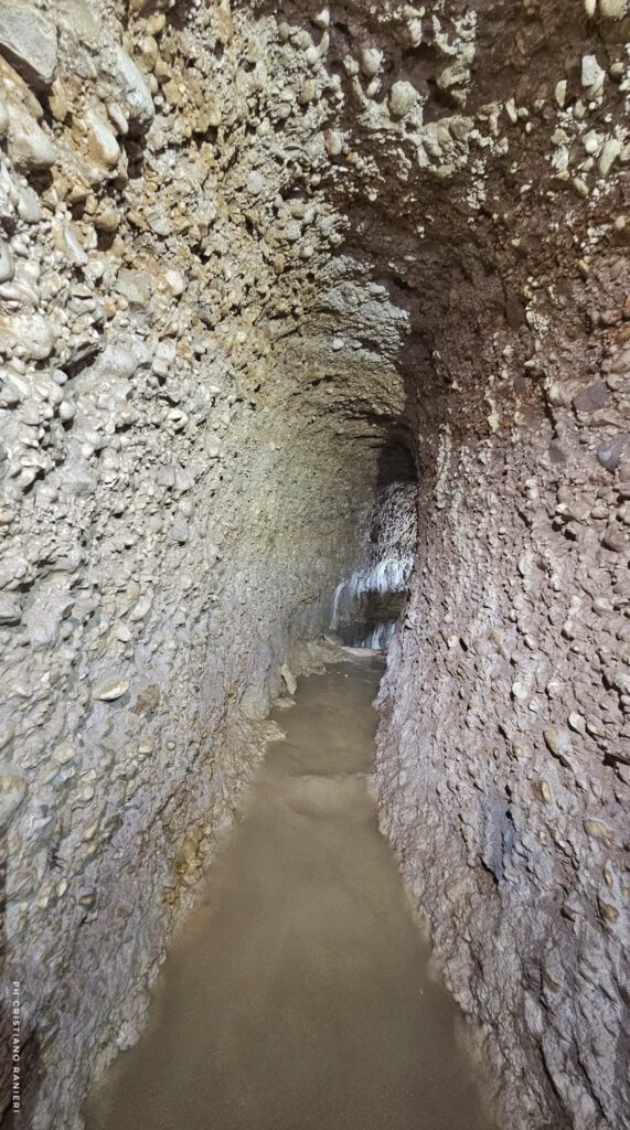 Narrow underground water channel with rough rock walls and a shallow pool of water inside the pre-Roman aqueduct in Italy.