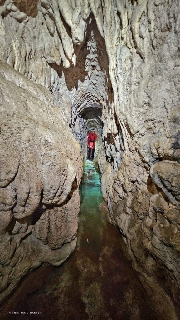Researcher in red protective clothing standing inside a long, narrow, rock-cut aqueduct with water covering the floor.