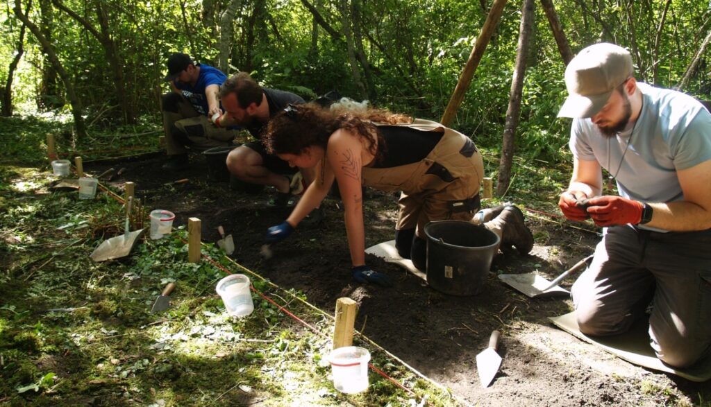 Archaeologists excavating a dark soil trench in a wooded area on Cepurīte Island during the Lake Indzers fieldwork in Latvia.