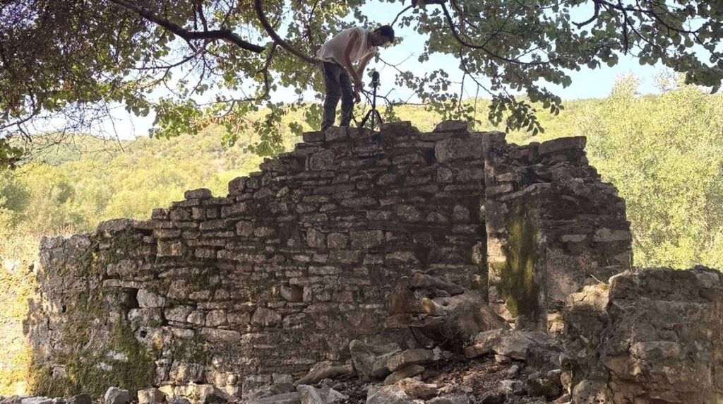 Archaeologist documenting a partially preserved stone building surrounded by vegetation at Butrint, Albania.