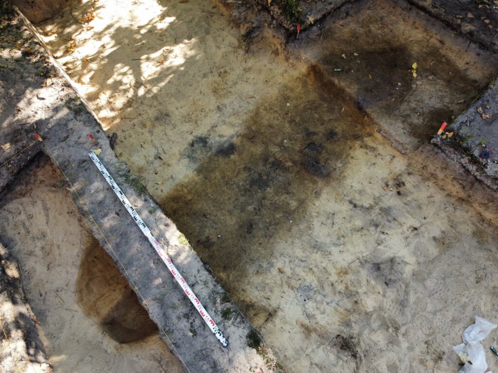 Excavation trench at Chodlik showing dark soil features, measuring scales, and the outline of a possible boat burial.