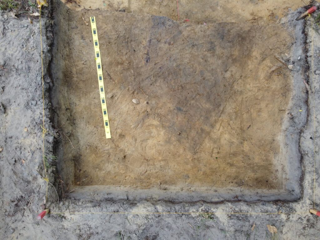 Rectangular archaeological excavation with a measuring scale and darker soil traces marking the investigated burial area.