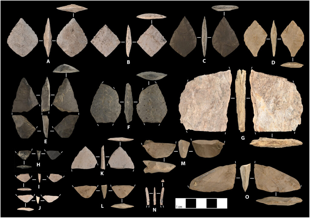 Photograph of bifacial stone tools and fragments from the Shég’ Xdaltth’í’ site in Alaska, including diamond-shaped bifaces, rough and finished bifaces, preforms, and a burin spall, arranged and labeled by letter.