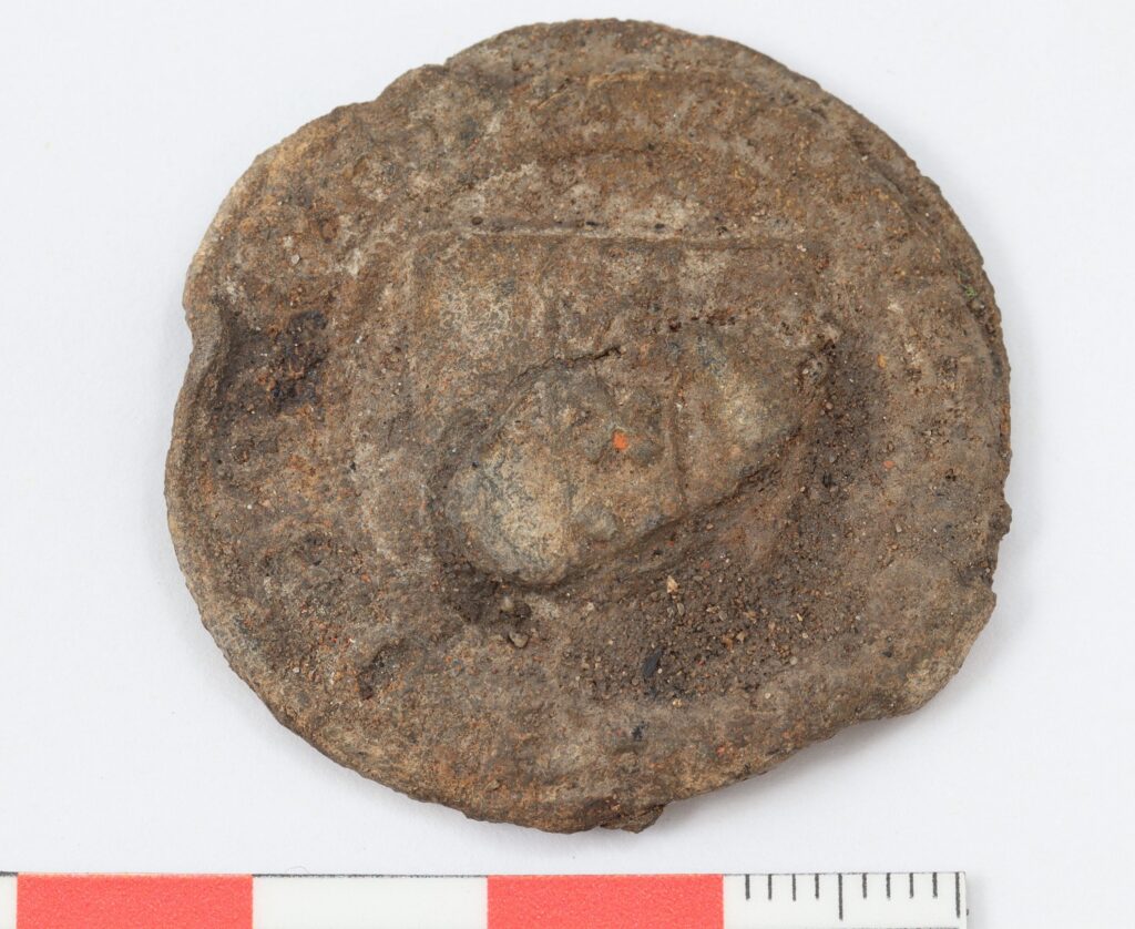 Reverse side of a weathered medieval lead cloth seal from Amsterdam, discovered in Tartu, Estonia, with a measurement scale beneath it.