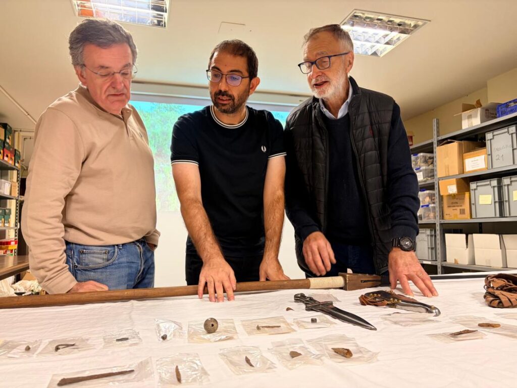 
Three researchers examining archaeological artifacts, including metal objects and tools, displayed on a table during the La Peña del Castro investigation.