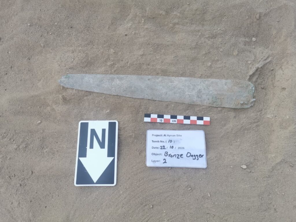 Ancient bronze dagger documented with an archaeological identification label and measuring scale during excavations at the Al Aynain burial site in Oman.