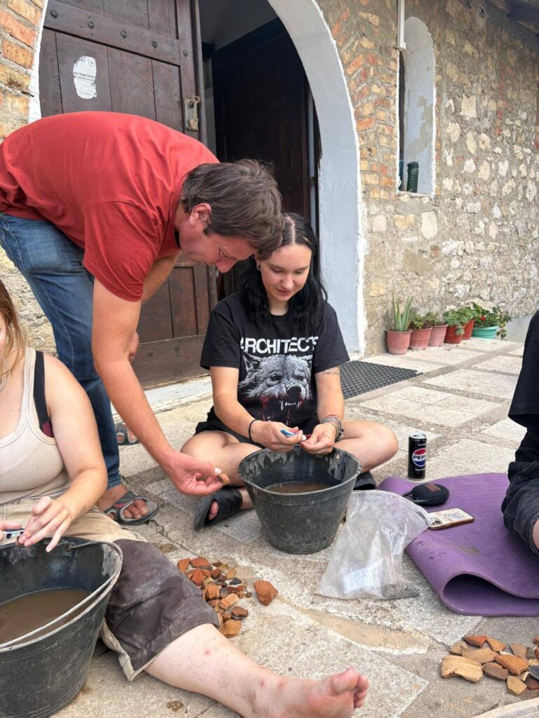 Archaeologists cleaning ancient pottery fragments beside buckets of water, with additional ceramic pieces arranged on the ground.