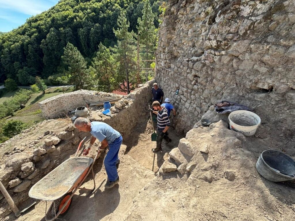 Archaeologists excavating beside the stone walls of medieval Teočak Fortress in Bosnia and Herzegovina, with a steep wooded hillside and road visible below.